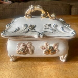 May include: A white porcelain trinket box with a gold handle and floral designs. The box has four feet and is decorated with gold accents.