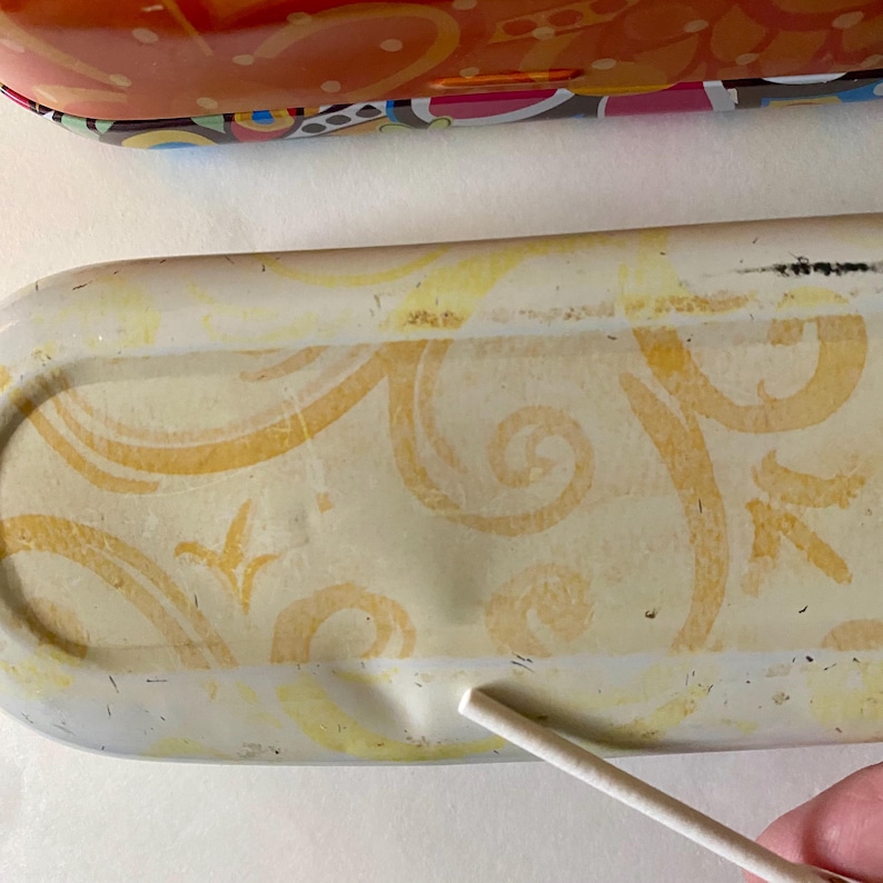 May include: Close-up of a decorative, rectangular metal tray with a cream-colored background and yellow floral design. The tray has a raised edge and a small indentation. A portion of a colorful tray is visible in the background.