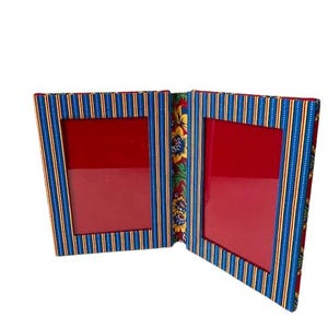 May include: A double photo frame with a red interior and a striped blue and yellow exterior. The frame is hinged in the middle and has a floral pattern on the spine.