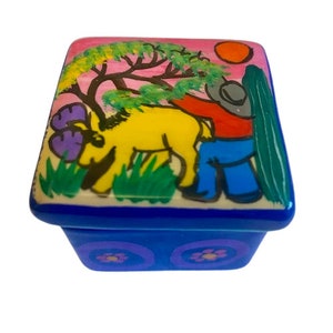 Peut inclure: Une petite boîte à bijoux carrée en céramique avec un motif peint à la main coloré. Le couvercle présente un animal jaune avec une corne violette, une personne en chemise rouge avec un chapeau bleu et des arbres verts sur un fond rose. La boîte a une base bleue avec des motifs floraux violets et blancs.
