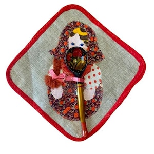Puede incluir: Un salvamanteles acolchado decorativo con un borde cosido rojo, con un diseño de muñeca Matryoshka. Una cuchara de madera pintada a mano con un diseño floral descansa sobre la muñeca. La cuchara tiene un mango dorado y una punta roja.