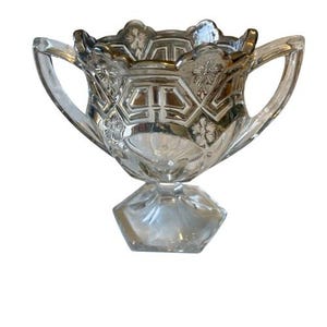 May include: A clear glass bowl with two handles and a decorative floral and geometric pattern. The bowl sits on a hexagonal base. The rim of the bowl has a scalloped edge.