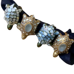 Pode incluir: Quatro argolas de guardanapo decorativas em forma de tartaruga. As argolas são feitas de um mosaico de azulejos verdes, dourados e brancos iridescentes. As tartarugas estão dispostas em fila sobre um fundo de tecido preto.