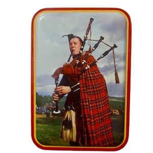 May include: A rectangular tin featuring a bagpiper in traditional Scottish attire. The bagpiper wears a red and black plaid kilt and plays the bagpipes. The background shows a green field, cloudy sky, and vintage cars.