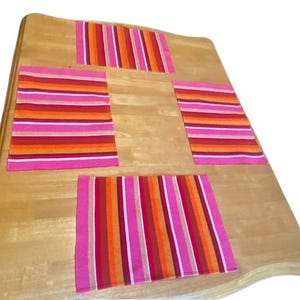 May include: A wooden table with four rectangular placemats and one smaller runner. The placemats and runner feature horizontal stripes in shades of pink, orange, red, and white. The table has a light brown finish.
