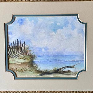 Puede incluir: Acuarela de una escena de playa con un océano azul, cielo y nubes. Una valla de madera y hierba de duna están a la izquierda. La pintura está enmarcada con un passe-partout blanco y un borde turquesa.