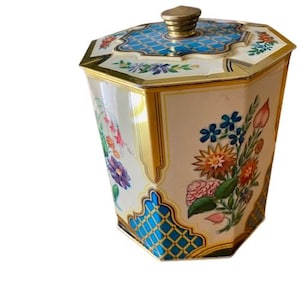 Puede incluir: Una lata octogonal antigua con tapa. La lata es de color crema con ribetes dorados y presenta diseños florales en tonos rosa, azul, naranja y morado. La tapa tiene un patrón geométrico azul y blanco y un pomo dorado.