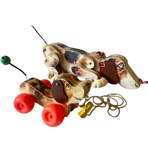 Op de afbeelding: Vintage houten trekdier speelgoed met rode wielen, een groene bal antenne en een bruin en wit gevlekt lichaam. De hond heeft een bruin oor en een rode halsband. Er is ook een ander hondenspeelgoed aanwezig.