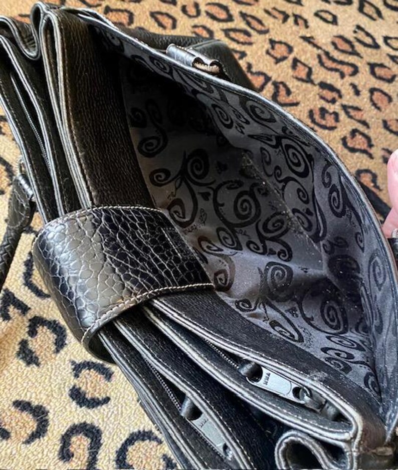 K&ouml;nnte beinhalten: Schwarze Ledertasche mit Rei&szlig;verschluss und gemustertem Futter. Die Tasche hat einen oberen Griff und einen abnehmbaren Schultergurt.