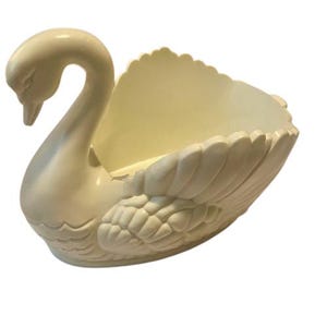 May include: A cream-coloured swan-shaped decorative bowl. The swan has a long, curved neck and a detailed wing design. The bowl is open at the top, creating a functional and decorative piece.