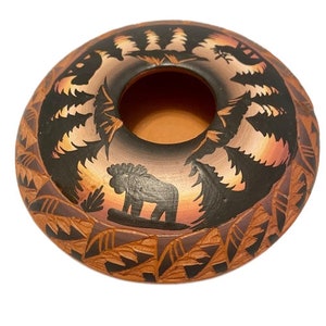 以下が含まれることがあります： 黒と白のデザインが施された、小さく丸い茶色の粘土製の壺。壺の中央には穴があり、木々と動物のシルエットで飾られています。