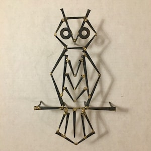 Op de afbeelding: Een metalen uilensculptuur gemaakt van spijkers. De uil is zwart met gouden accenten en heeft grote ronde ogen.