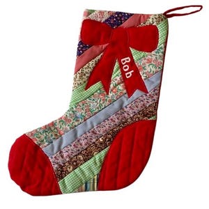 Puede incluir: Un calcetín navideño acolchado con un lazo rojo y el nombre "Bob" bordado. El calcetín presenta un diseño de patchwork con varias telas florales y estampadas en tonos rojos, verdes, azules y marrones. Se adjunta un lazo rojo para colgar.