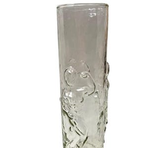 May include: A clear glass vase with a cylindrical form. The vase has a textured, embossed design of figures. The glass is transparent, allowing light to pass through. The vase is suitable for displaying flowers or as a decorative item.