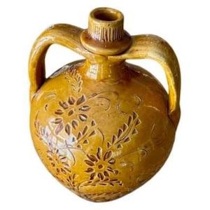 May include: A yellow-brown ceramic jug with two handles and a textured neck. The jug features floral and leaf patterns carved into the surface. The jug is a decorative item.
