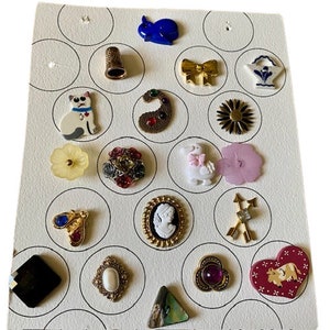 May include: A white card with 18 different buttons arranged in a grid pattern. The buttons are various colors and shapes, including a cat, a bunny, a flower, a heart, a swan, a bow, a thimble, and a paisley design.