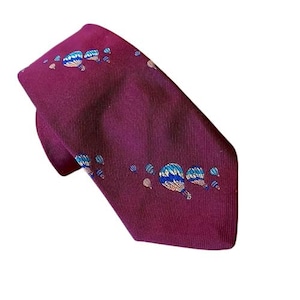 May include: A burgundy tie with a repeating pattern of blue, white, and gold hot air balloons. The tie is made of a ribbed fabric and is angled diagonally across the frame.