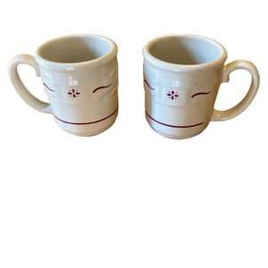 Può includere: Due tazze in ceramica color bianco sporco con una trama a intreccio in rilievo sulla metà superiore. Ogni tazza è decorata con un piccolo motivo floreale bordeaux e una sottile linea orizzontale bordeaux che circonda la parte inferiore.