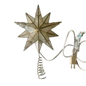 May include: A multi-pointed star tree topper featuring translucent white panels framed in gold-toned metal with delicate wire scrollwork accents. The star is attached to a coiled metal base and includes a white electrical cord with a plug.