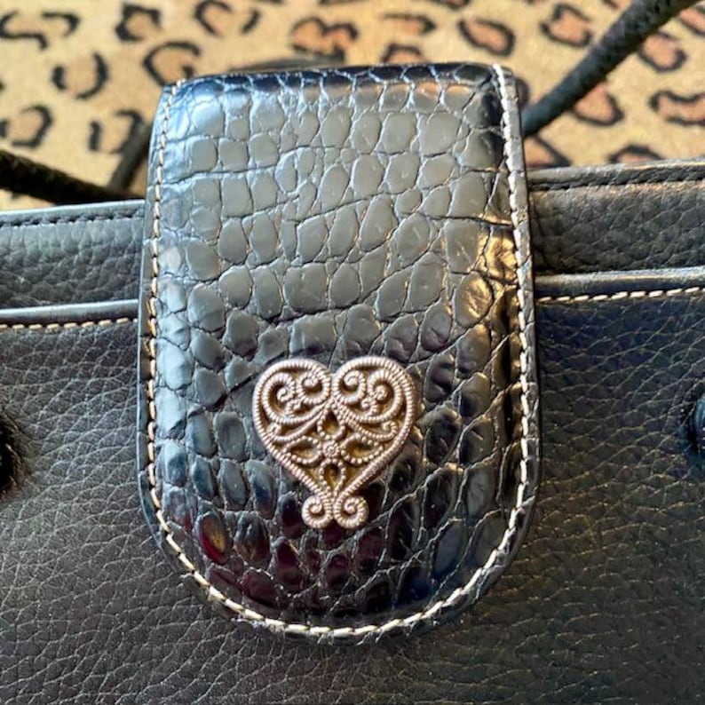 K&ouml;nnte beinhalten: Schwarze Lederb&ouml;rse mit einem herzf&ouml;rmigen Metallakzent. Die B&ouml;rse hat einen Klappverschluss und eine strukturierte Oberfl&auml;che.