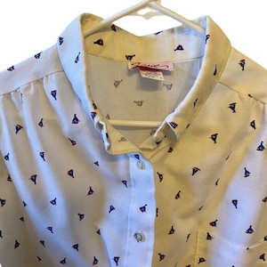 Op de afbeelding: Een wit overhemd met knopen en een klein zeilbootprint in blauw, rood en wit. Het shirt heeft een kraag en een knoopsluiting. Het label op het shirt luidt "Apparel".