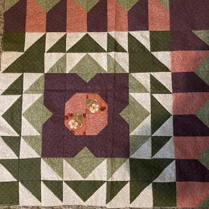 May include: A quilt with a geometric pattern of green, pink, and purple triangles. The center of the quilt features a flower-shaped design with pink and white floral fabric.