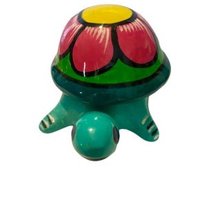 May include: A hand-painted ceramic turtle figurine. The shell features a pink flower design with black outlines and a yellow centre. The body is turquoise with black detailing.