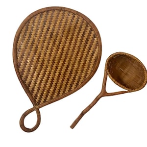 May include: Two woven rattan items. One is a large, heart-shaped fan with a looped handle. The other is a small, round basket attached to a wooden handle. Both items are in shades of brown and tan.