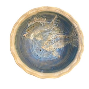 May include: A ceramic bowl with a blue interior and a beige rim. Two fish are sculpted in the center of the bowl. The fish are light beige with detailed fins and scales. The bowl has a wavy edge.