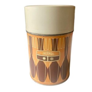 May include: Vintage Thermos brand insulated container. The container has a cream-colored lid and a cylindrical body with an orange and brown geometric design. The word "THERMOS" is printed on the front, along with a sun and snowflake symbol.