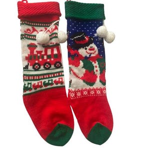 Può includere: Due calze natalizie in maglia con pon pon bianchi. La calza a sinistra è rossa e bianca con un motivo a trenino rosso. La calza a destra è blu, bianca e rossa con un pupazzo di neve che indossa un cilindro nero. Entrambe hanno polsini colorati.