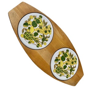 Op de afbeelding: Een houten dienblad met twee witte keramische tegels. De tegels hebben een groen en gouden bloemenmotief met fruit en bladeren.