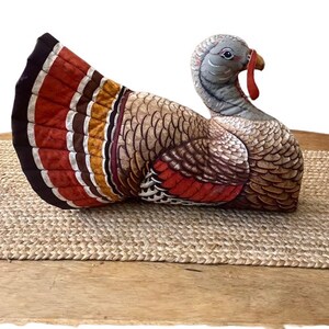 Puede incluir: Una figurita de pavo de peluche con un cuerpo estampado en marrón, naranja y blanco y una barbilla roja. El pavo está tumbado de lado con las plumas de la cola extendidas.