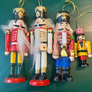 May include: A collection of four wooden nutcracker ornaments. The ornaments feature detailed painted faces, uniforms, and hats in red, white, yellow, and blue. Each ornament has a gold string for hanging, perfect for holiday decor.