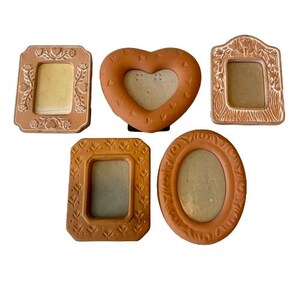 May include: Five terracotta-colored picture frames in various shapes: rectangular, heart-shaped, and oval. The frames have decorative embossed floral and geometric patterns. The heart-shaped frame has a cutout center.