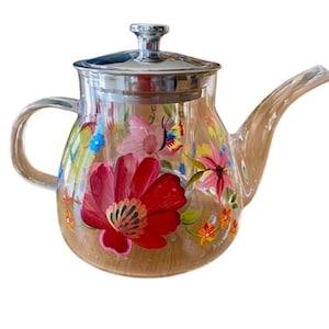Könnte beinhalten: Eine klare Glasteekanne mit einem Edelstahldeckel und einem floralen Design, das auf die Seite gemalt ist. Die Blumen sind rosa, rot, gelb und blau, und auf der Seite ist ein Schmetterling gemalt.