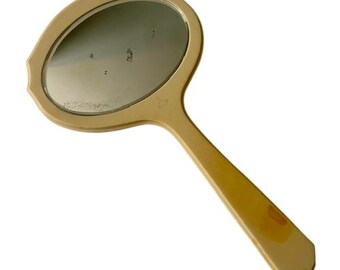 May include: A vintage hand mirror with an oval-shaped reflective surface and a long, slender handle. The mirror has a light yellow frame and handle, with a slightly ornate edge. The handle has a small, octagonal end. The mirror is likely made of plastic.