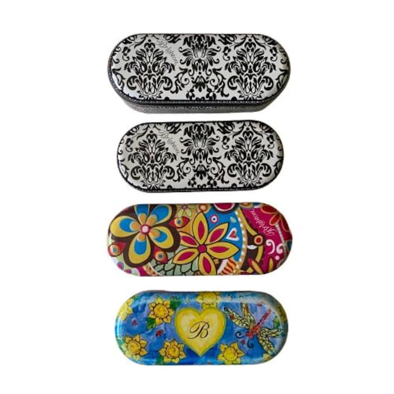 May include: Four oval-shaped eyeglass cases. Two cases feature a black and white damask pattern. The other two cases have colorful floral and nature-themed designs. One case has a yellow heart with the letter "B".