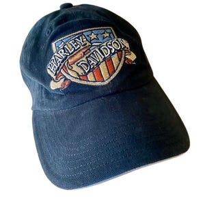 Può includere: Un cappellino da baseball blu navy con un logo Harley-Davidson ricamato. Il logo è un design a scudo con il nome del marchio in un banner, che incorpora colori rosso, bianco e blu. Il cappellino ha una visiera curva e un bottone superiore.