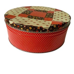 May include: A round, red tin with a white polka dot pattern on the sides. The lid features a patchwork design with red, black, and cream-colored squares. The tin is likely used for storage or as a decorative item.