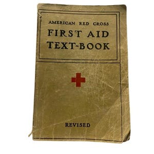 Puede incluir: Un libro de bolsillo vintage de color beige titulado American Red Cross First Aid Text-Book con una cruz roja en el centro y la palabra Revised impresa cerca de la parte inferior. La cubierta muestra signos de desgaste con pliegues visibles.