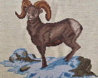 May include: A cross-stitch depiction of a ram with large, curved brown horns. The ram is brown and stands on a snowy, blue and white landscape. The background is a neutral beige color.