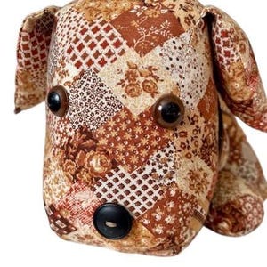 May include: A handmade dog-shaped plush toy. The toy is made from various fabric patches in shades of brown, beige, and cream, with floral and geometric patterns. It has large, brown button eyes and a black button nose. The ears are floppy.