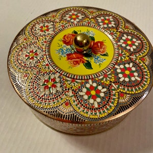 Op de afbeelding: Een ronde, vintage blikken koekjesbus met een geel deksel met een bloemenmotief van rode rozen, blauwe bloemen en groene bladeren. De bus is versierd met een kleurrijk, ingewikkeld patroon van witte, rode, gele, blauwe en groene stippen en swirls.