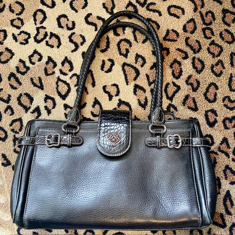 K&ouml;nnte beinhalten: Schwarze Ledertasche mit einem herzf&ouml;rmigen Logo und zwei Schnallen auf der Vorderseite. Die Tasche hat einen Henkel und einen Schultergurt.