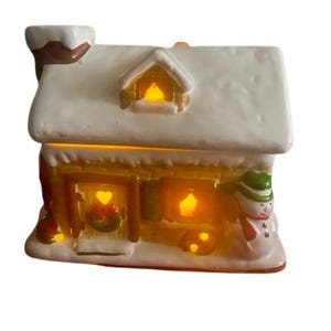 Op de afbeelding: Een kerst huis van keramiek met een sneeuwpop en verlichte ramen. Het huis is versierd met sneeuw en een krans op de deur.