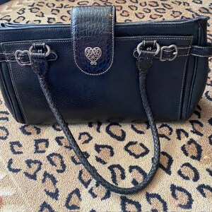 K&ouml;nnte beinhalten: Eine schwarze Ledertasche mit einem geflochtenen Riemen und einem silbernen herzf&ouml;rmigen Anh&auml;nger. Die Tasche hat einen Klappverschluss und einen oberen Griff.