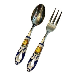 May include: A stainless steel spoon and a three-tined fork featuring ceramic handles. The handles are white with a hand-painted yellow lemon motif, green leaves, and blue scrollwork accents, ending in a circular cutout at the base of each.