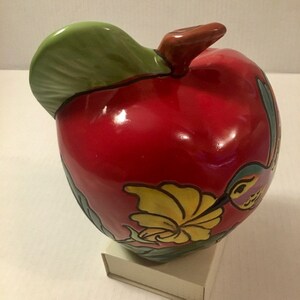 May include: A red ceramic apple with a green leaf and a hummingbird painted on the side. The apple is decorated with yellow and green flowers.