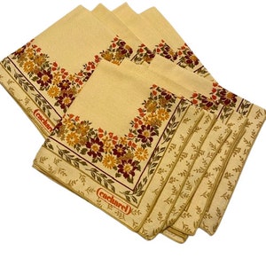 May include: Set of six beige linen napkins with a floral border in shades of red, orange, and yellow. The napkins have a "Cacharel" logo in orange.